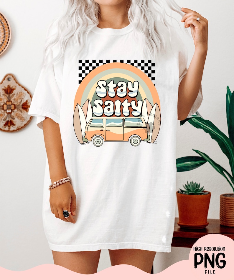 May include: A white t-shirt with a graphic design of a vintage van driving past surfboards with a rainbow and the text "Stay Salty" in a retro font.
