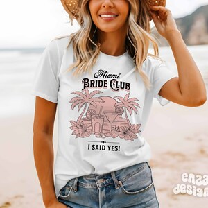 May include: White t-shirt with a pink graphic design that says "Miami Bride Club" with palm trees, a sunset, and cocktails. The text "I said yes!" is below the graphic.