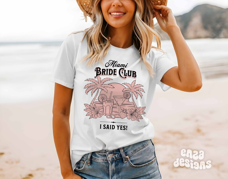 May include: White t-shirt with a pink graphic design that says "Miami Bride Club" with palm trees, a sunset, and cocktails. The text "I said yes!" is below the graphic.