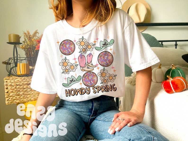 May include: White t-shirt with a colorful graphic design featuring cowboy boots, disco balls, daisies, and cowboy hats. The text "HOWDY SPRING" is printed below the graphic. The shirt has a relaxed fit.