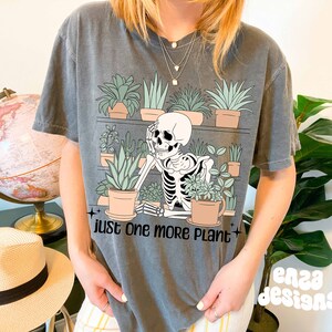 May include: A gray t-shirt with a skeleton holding a potted plant. The shirt has the text "Just one more plant" printed on it.