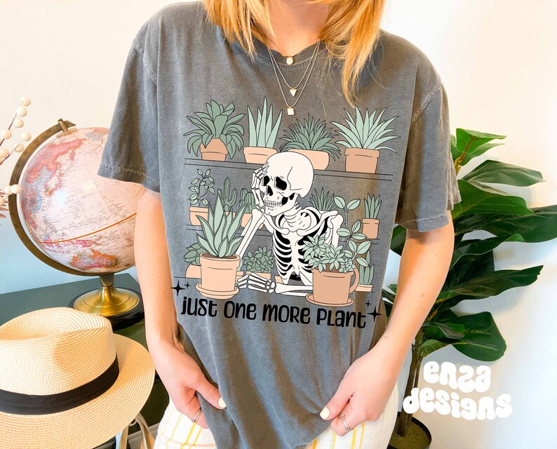 May include: A gray t-shirt with a skeleton holding a potted plant. The shirt has the text "Just one more plant" printed on it.