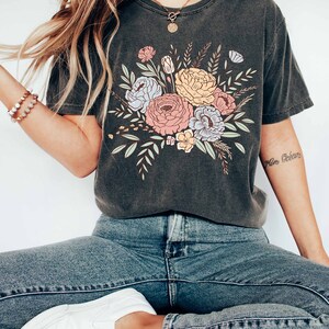 May include: A dark gray t-shirt with a floral print featuring pink, yellow, blue, and green flowers. The shirt has a crew neck and short sleeves. The text "More B&eacute;b&eacute;" is visible on the right sleeve.