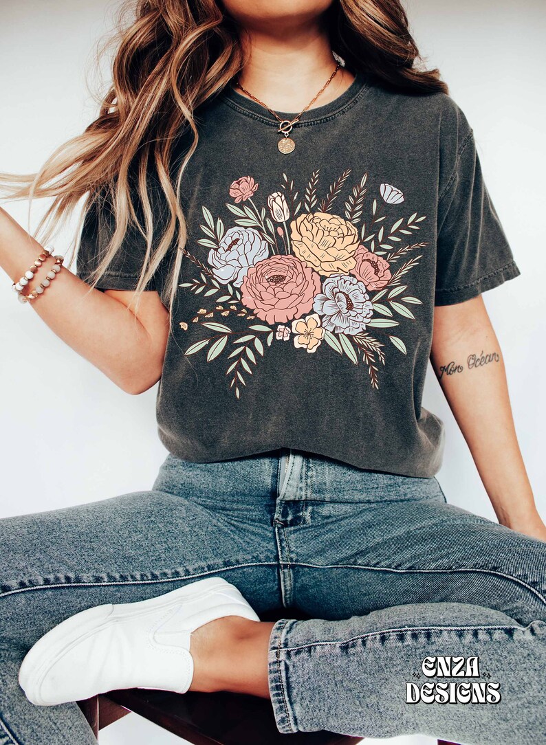 May include: A dark gray t-shirt with a floral print featuring pink, yellow, blue, and green flowers. The shirt has a crew neck and short sleeves. The text "More B&eacute;b&eacute;" is visible on the right sleeve.
