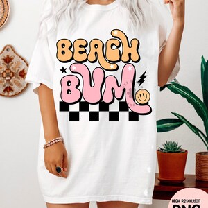 May include: White t-shirt featuring a graphic design that reads "Beach Bum" in pink and orange bubble letters with a black and white chequered pattern underneath. The graphic also includes a smiley face and a lightning bolt.