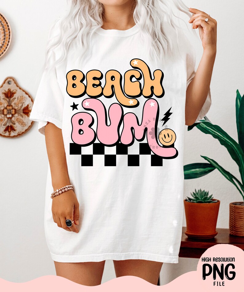May include: White t-shirt featuring a graphic design that reads "Beach Bum" in pink and orange bubble letters with a black and white chequered pattern underneath. The graphic also includes a smiley face and a lightning bolt.