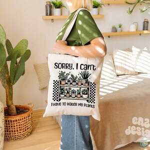 Puede incluir: Una bolsa de lona blanca con un dise&ntilde;o a cuadros blanco y negro. La bolsa presenta un dibujo de un estante con plantas en macetas y el texto "Lo siento, no puedo, tengo que regar mis plantas".