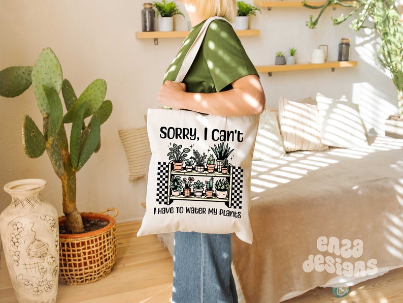 Puede incluir: Una bolsa de lona blanca con un dise&ntilde;o a cuadros blanco y negro. La bolsa presenta un dibujo de un estante con plantas en macetas y el texto "Lo siento, no puedo, tengo que regar mis plantas".