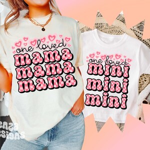 May include: Two white t-shirts with pink checkered text. The larger shirt says "one loved mama mama mama" and the smaller shirt says "one loved mini mini mini".