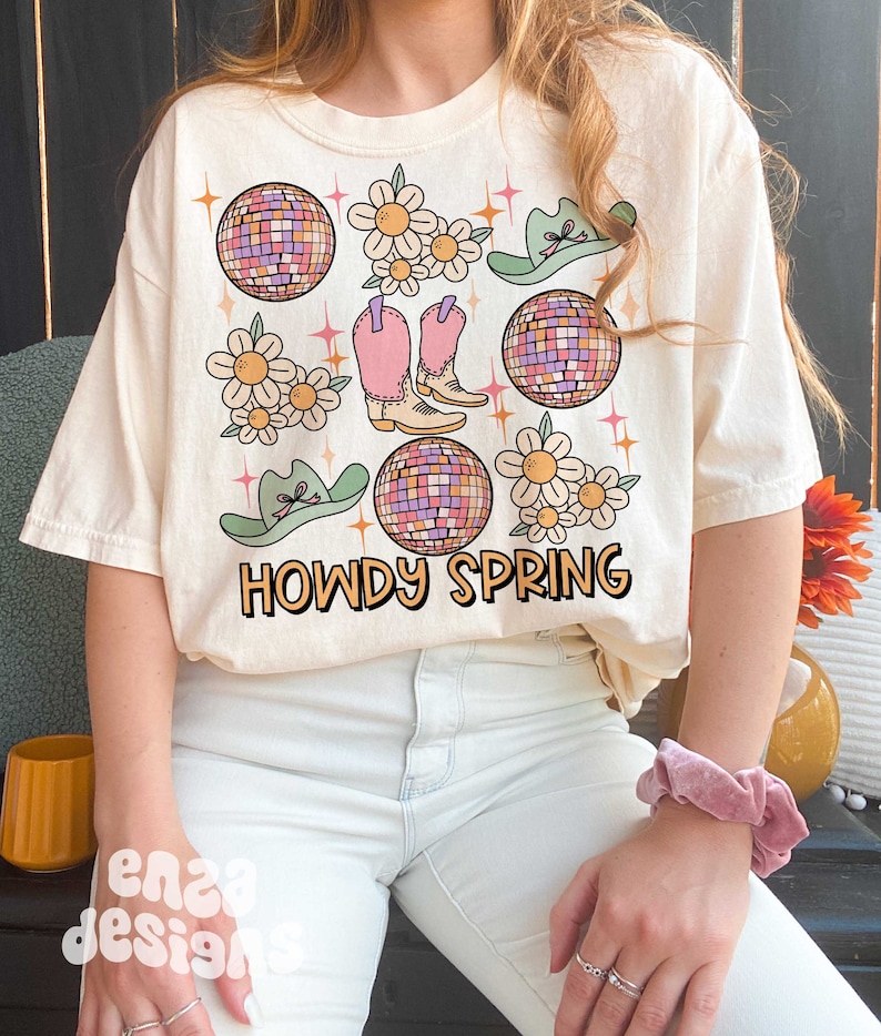 May include: Cream-colored t-shirt with a colorful graphic design. The design includes disco balls, flowers, cowboy boots, and hats. The text "HOWDY SPRING" is printed in orange and black. The shirt is a relaxed fit.