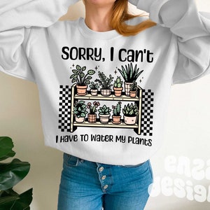 Puede incluir: Sudadera blanca con el texto "Lo siento, no puedo, tengo que regar mis plantas" y un gr&aacute;fico de una estanter&iacute;a con plantas en macetas.