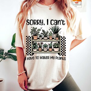 Puede incluir: Una camiseta blanca con el texto "Lo siento, no puedo, tengo que regar mis plantas" y una ilustraci&oacute;n de dibujos animados de un estante con plantas en macetas.