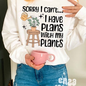 May include: White sweatshirt with the text "Sorry I can't... I have plans with my plants" and a cartoon illustration of a potted plant on a stool with a watering can.