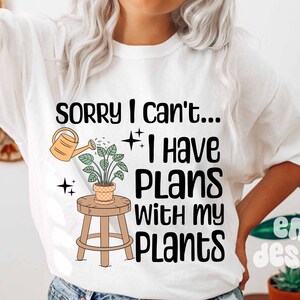 May include: White t-shirt with the text "Sorry I can't... I have plans with my plants."  A cartoon illustration of a potted plant with a watering can pouring water on it is above the text.