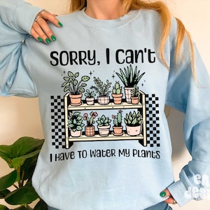 Puede incluir: Sudadera azul claro con un gr&aacute;fico de una estanter&iacute;a de plantas con el texto "Lo siento, no puedo, tengo que regar mis plantas".