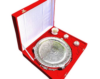 May include: A silver-plated puja thali set with an Om symbol in the center, surrounded by intricate designs. The set comes in a red velvet box with a white interior.