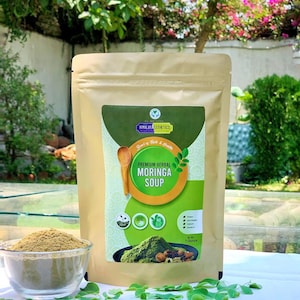 May include: A brown paper bag with a green and yellow label that says "Premium Herbal Moringa Soup" and "Himalaya Aesthetics". The bag contains a green powder and is sitting on a white surface with green leaves.
