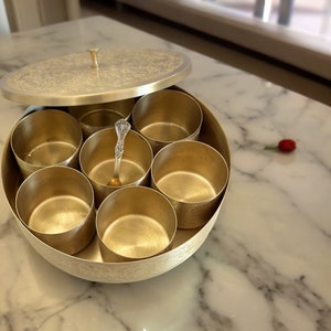 May include: A silver-colored spice box with a lid, featuring an intricate floral design. The box is divided into seven round compartments, each holding a small, gold-colored cup. A decorative spoon rests inside. The box sits on a marble surface.