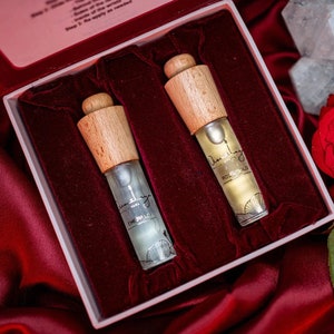 May include: Two small glass bottles with wooden caps and gold labels. The bottles are in a red velvet lined box. The labels read "Emerald" and "Rose Gold".