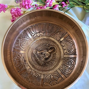 May include: A copper singing bowl with intricate embossed designs, featuring a small Buddha figure in the center. The bowl has a smooth, polished surface and a slightly flared rim.