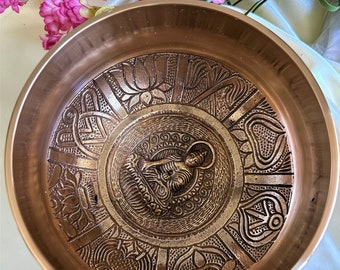 Puede incluir: Un cuenco de canto de cobre con intrincados diseños en relieve, que presenta una pequeña figura de Buda en el centro. El cuenco tiene una superficie lisa y pulida y un borde ligeramente acampanado.