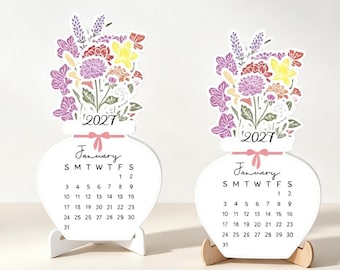 May include: Two white vase-shaped desk calendars for January 2027, each featuring a colorful floral illustration of purple, red, and yellow flowers at the top, a pink ribbon graphic, and a standard monthly grid with the days of the week listed.