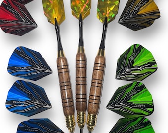 May include: Three gold darts with black shafts and wooden barrels. The darts are surrounded by a variety of blue, green, and red dart flights with the word "Harrows" and "Supergrip Ultra" printed on them.