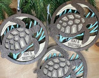 May include: Three wooden ornaments shaped like sea turtles with blue and white accents. Each ornament has the text "Seas & Greetings" on it.