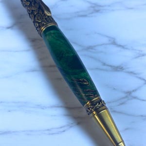 Op de afbeelding: Een handgemaakte pen met een groene gemarmerde schacht en sierlijke goudkleurige accenten. De pen heeft een gedetailleerd bloemmotief aan de bovenkant en decoratieve banden. De punt van de pen heeft een klassieke conische vorm.
