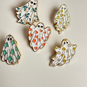 May include: Five white ghost pins with different patterns. The ghosts are wearing sunglasses, hearts, flowers, and smiley faces. The pins are gold-toned.