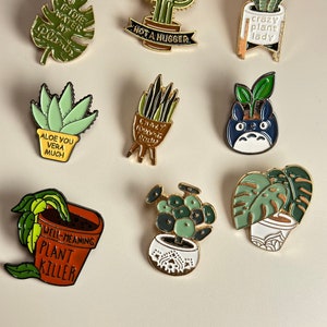 May include: Set of ten enamel pins featuring various plants and puns. The pins are gold-toned with green, white, and brown accents. The pins feature phrases like "If I Die Water My Plants", "Not a Hugger", "Crazy Plant Lady", "Aloe You Vera Much", "Crazy Plant Mom", "Well-Meaning Plant Killer", and more.