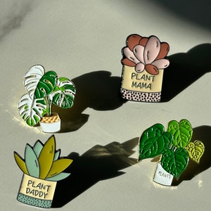May include: Four enamel pins featuring plant designs. One pin shows a monstera plant, another says "PLANT MAMA", and a third says "PLANT DADDY". The fourth pin says "PLANTS". The pins have a white, green, yellow, and brown color scheme.