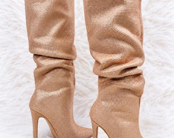 May include: A pair of knee-high boots in a shimmering rose gold tone, featuring a pointed toe and a slender stiletto heel. The exterior surface is covered in a dense, fine-textured grid of small reflective studs, with a slouchy shaft design.