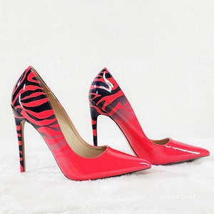 May include: A pair of pointed-toe high heel pumps featuring a glossy finish. The shoes display a vibrant red base color with a black zebra stripe pattern concentrated on the heel and back section. The text TWF SHOES is visible in the bottom right.