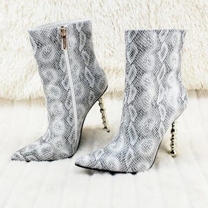 May include: A pair of ankle boots featuring a grey and white snakeskin pattern, a pointed toe, and a gold-toned zip on the side. The boots are elevated by a unique gold-toned heel constructed from a vertical stack of small, polished spheres.