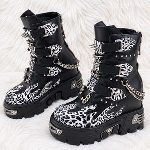 May include: A pair of black platform boots featuring white and black leopard print flame patterns on the toe and sides. The boots include multiple buckled straps, silver-toned metal spikes, decorative chains, and a thick lugged rubber sole.