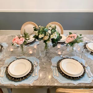 May include: A table setting with blue placemats, white plates, silver cutlery, and pink flowers in glass vases. The table is made of marble and has a gray and white pattern.
