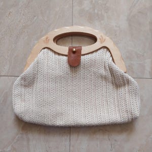 May include: Cream-colored woven handbag with a light wood handle. The handle has a curved design with a floral pattern etched on each side. A brown leather strap secures the bag's closure. The bag is sitting on a light-colored tile surface.