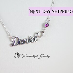 May include: Silver necklace with the name "Daniel" in cursive script, a small heart charm, and a single red gemstone. The necklace is on a white background.