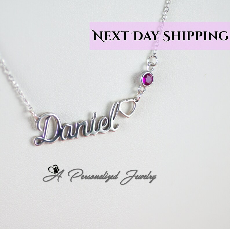 May include: Silver necklace with the name "Daniel" in cursive script, a small heart charm, and a single red gemstone. The necklace is on a white background.