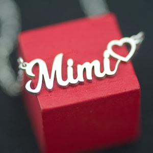 May include: Silver necklace with the name "Mimi" spelled out in cursive lettering with a small heart charm at the end. The necklace is displayed on a red box.