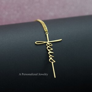 May include: A gold necklace with a cross pendant that has the word "Jesus" written in cursive on it.