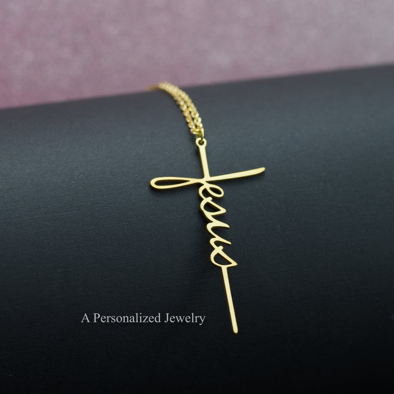 May include: A gold necklace with a cross pendant that has the word "Jesus" written in cursive on it.