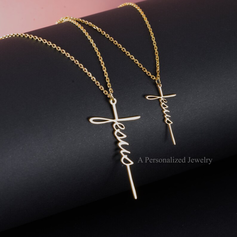 May include: Two gold necklaces with a cross pendant featuring the word "Jesus" in cursive. The necklaces are on a black background.