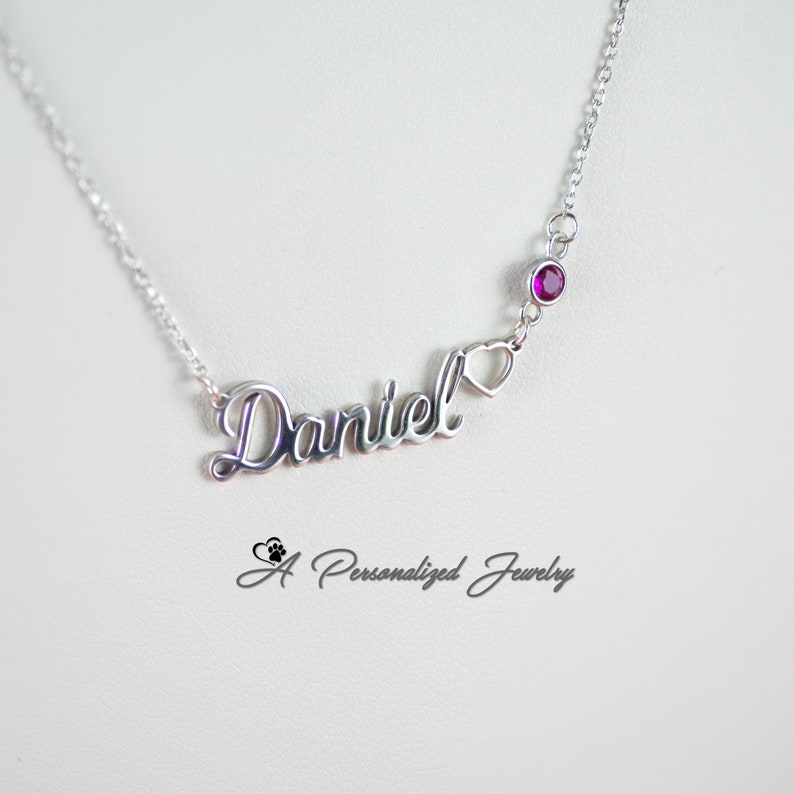 May include: Silver necklace with a cursive name "Daniel" and a small heart charm. A single purple gemstone is attached to the chain above the name.