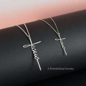 May include: Two silver necklaces with a cursive "Jesus" cross pendant. The necklaces are on a black surface with the text "A Personalized Jewelry" in the bottom right corner.