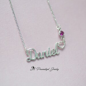May include: Silver necklace with the name "Daniel" spelled out in cursive script. A small heart charm hangs from the end of the name, with a pink gemstone above it.