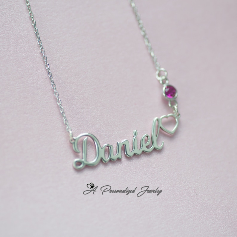 May include: Silver necklace with the name "Daniel" spelled out in cursive script. A small heart charm hangs from the end of the name, with a pink gemstone above it.