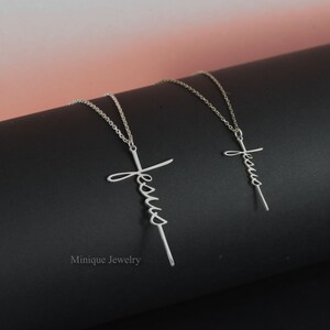 May include: Two silver necklaces with a cross pendant featuring the word "Jesus" in cursive script. The necklaces are on a black surface.
