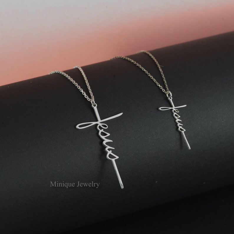 May include: Two silver necklaces with a cross pendant featuring the word "Jesus" in cursive script. The necklaces are on a black surface.